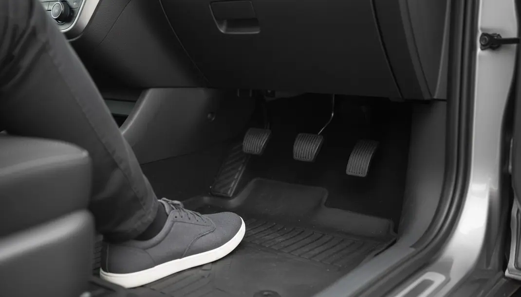 Pé esquerdo de um condutor pousado no piso do carro, ao lado do pedal da embraiagem, com os três pedais à vista