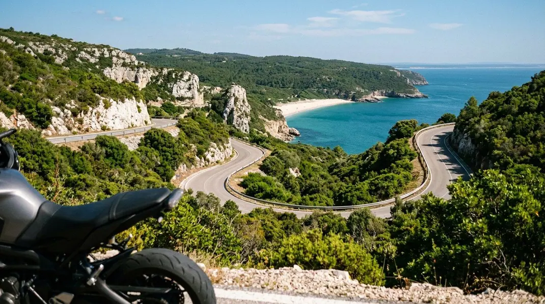 Estrada sinuosa a descer pela serra coberta de mato verde até uma baía de água turquesa com praia de areia branca
