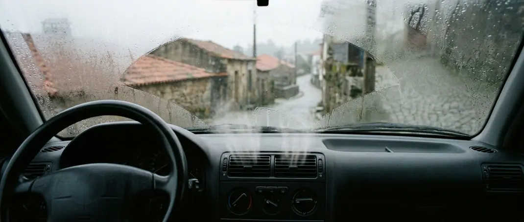 Para-brisas embaciado por dentro numa manhã de chuva, com o ar da ventilação a abrir uma zona limpa sobre o tablier